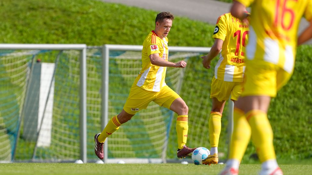 A player in yellow soccer gear dribbles the ball on a sports field, while other players in yellow jerseys stand in the background.