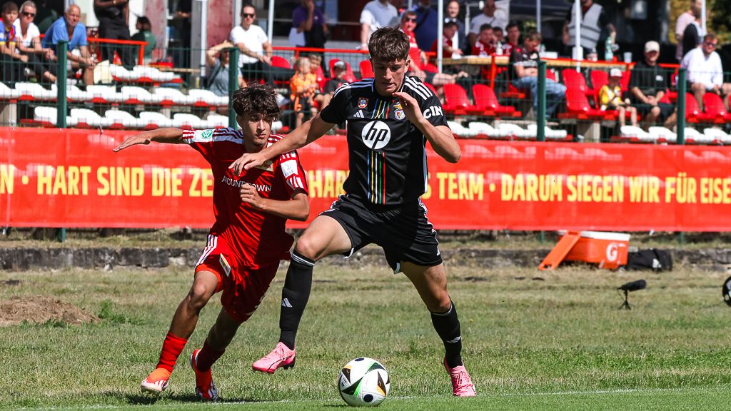 Ein Fußballspieler in schwarzem Trikot dribbelt den Ball, während ein Gegner in rotem Trikot ihn verfolgt. Zuschauer im Hintergrund.
