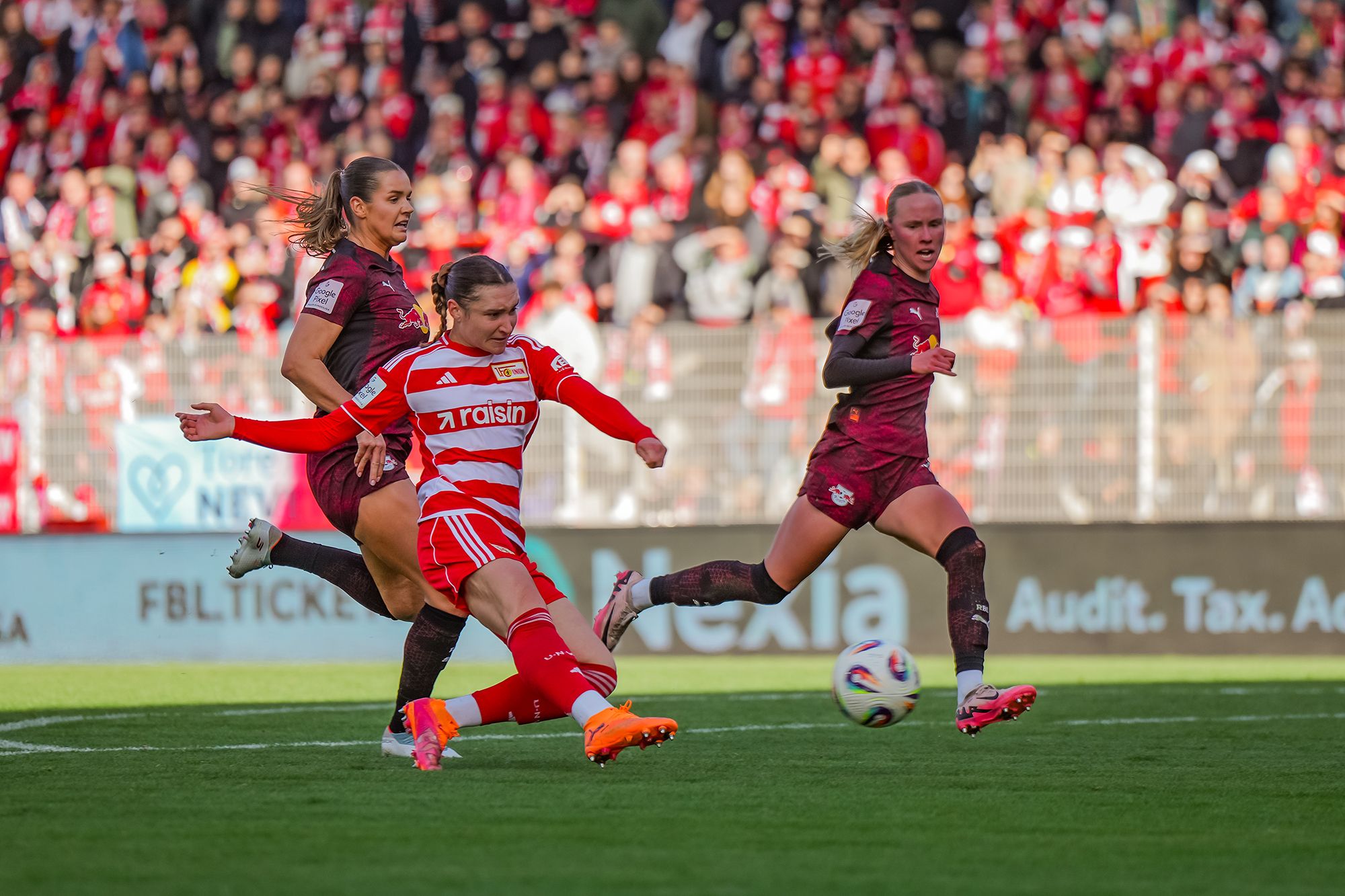 Spielszene im Fußball mit einer Spielerin in roter und weißer Kleidung, die den Ball schießt, während zwei Gegnerinnen in dunklerer Kleidung reagieren.
