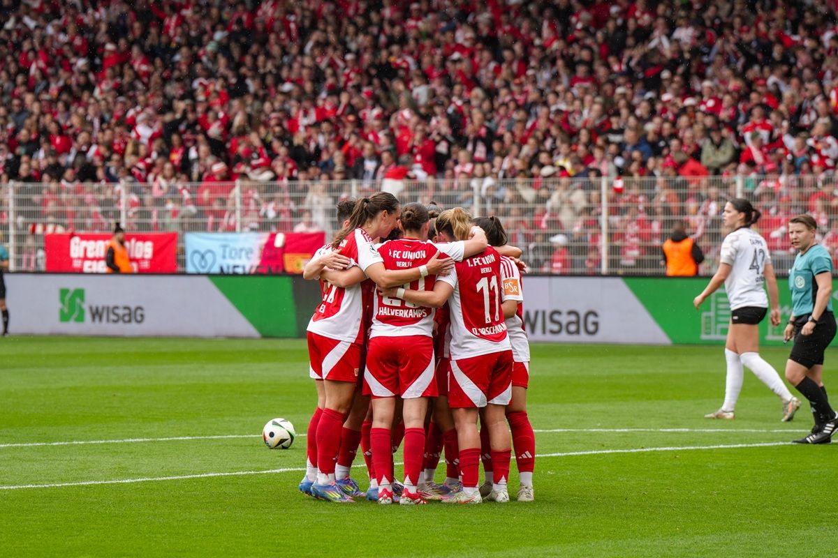 Mannschaft von Spielerinnen steht umarmt auf dem Spielfeld, jubelnd inmitten einer jubelnden Menge.