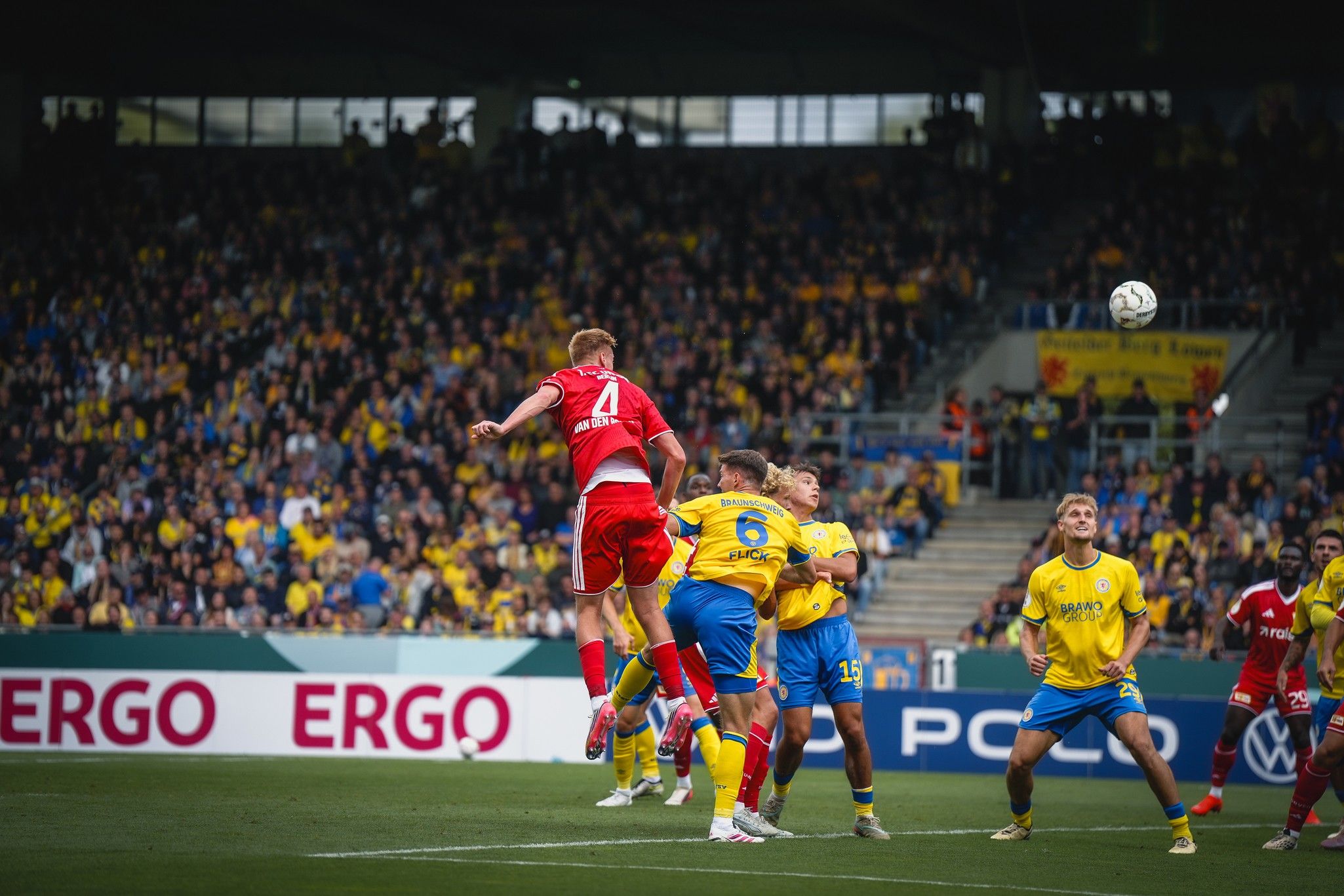 Ein Fußballspieler springt hoch, um den Ball bei einem Spiel zwischen zwei Mannschaften zu köpfen, während Zuschauer im Hintergrund stehen.