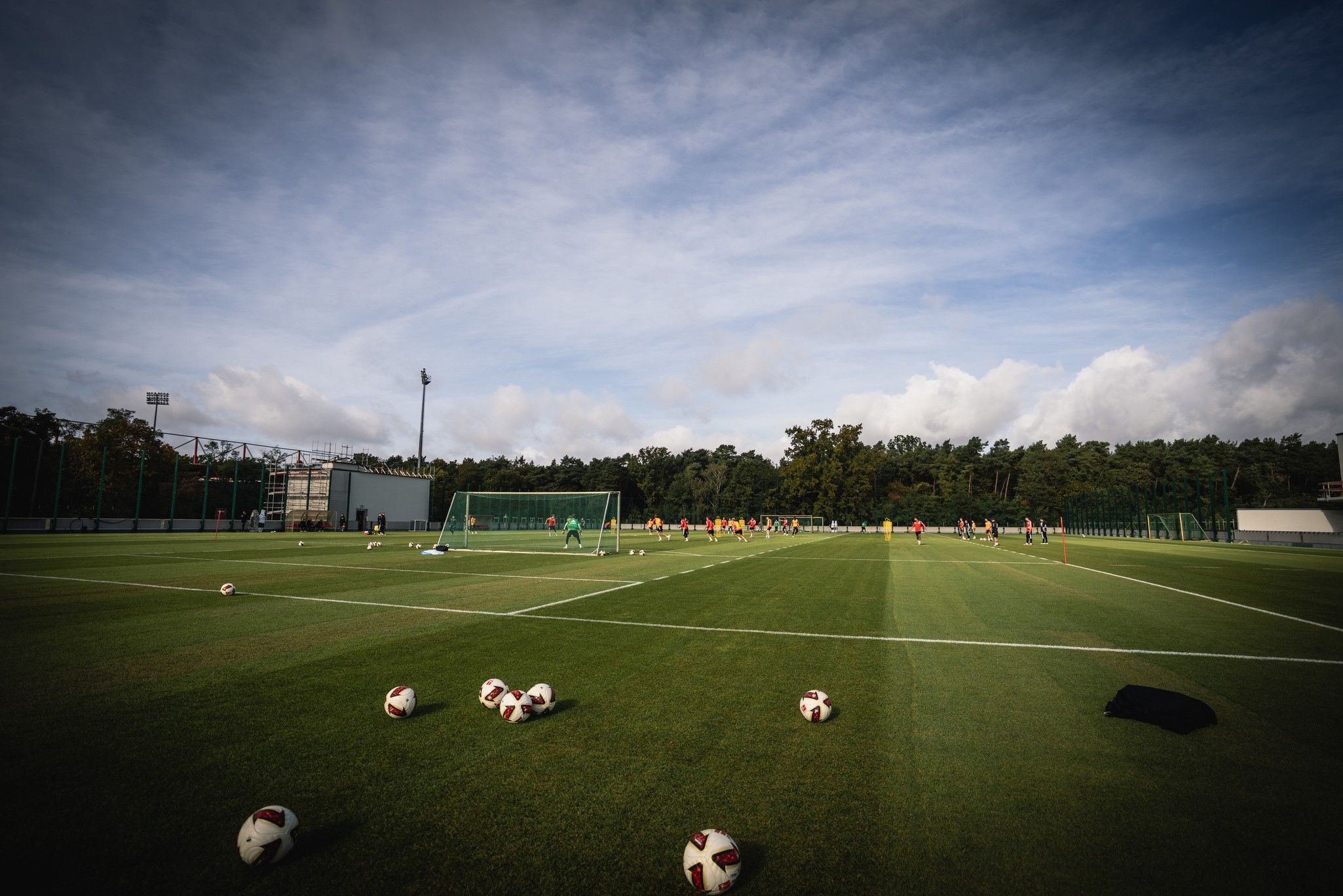 Fußballplatz mit mehreren Bällen, auf dem Spieler trainieren, umgeben von Bäumen und blauem Himmel.