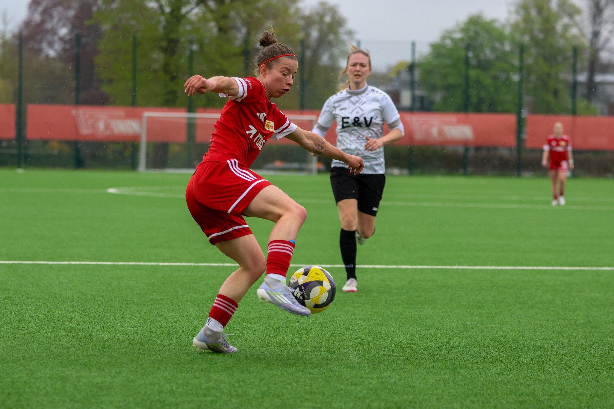 Spielerin in rotem Trikot führt einen Schuss auf dem Fußballfeld aus, während eine Gegnerin im Hintergrund zusieht.