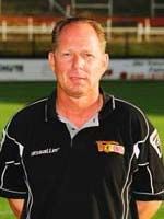 Ein Mann steht auf einem Fußballfeld, trägt ein schwarzes Polohemd mit weißen Akzenten und hat ein kurzes, blondes Haar.