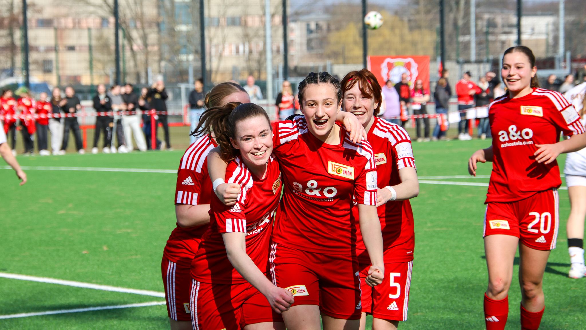 Jubilierende Fußballspielerinnen in roten Trikots feiern nach einem Tor auf einem Rasenplatz.