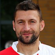 Ein Mann mit kurzen, dunklen Haaren und Bart lächelt in einem sportlichen, weißen Trikot.