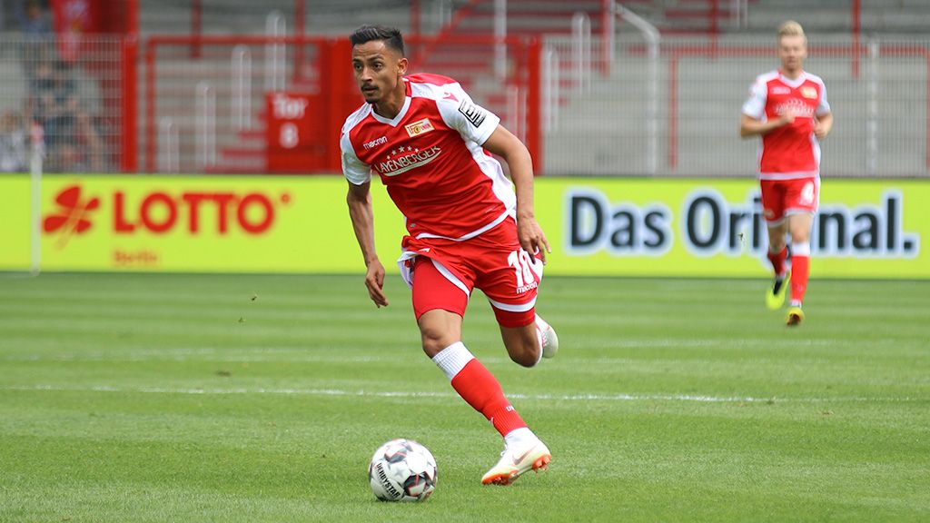 Spieler in roter Fußballuniform sprintet über das Spielfeld, während ein weiterer Spieler im Hintergrund steht.