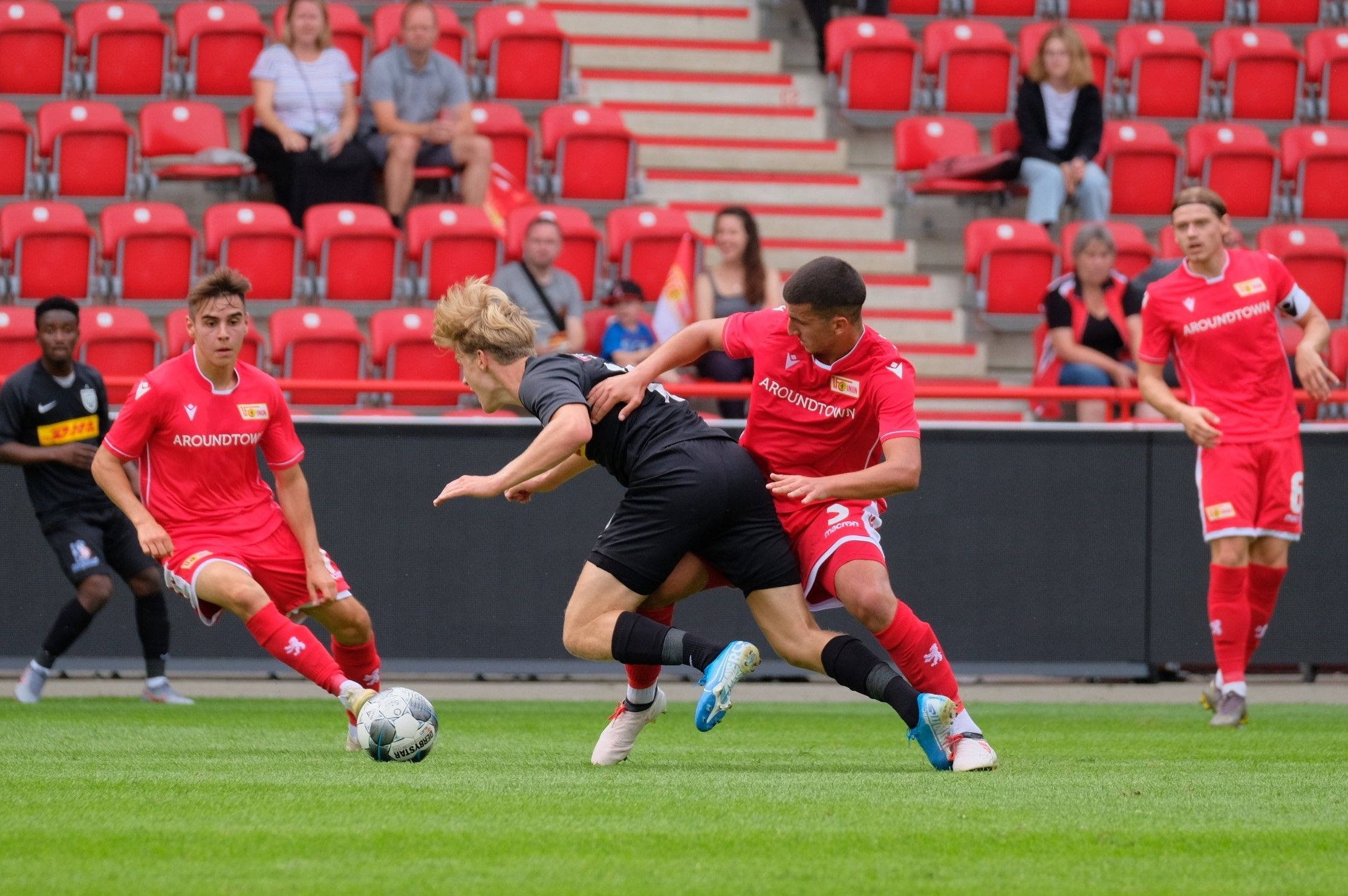 Zwei Fußballspieler in roten Trikots versuchen, einen Ball von einem Spieler in schwarzem Trikot zu erobern, während Zuschauer im Hintergrund sitzen.