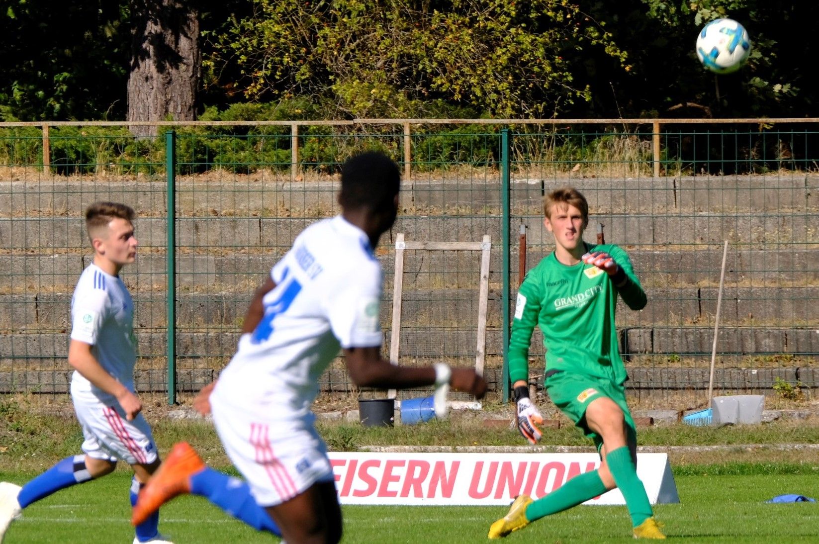 Ein Fußballtorwart fängt den Ball, während zwei Spieler auf dem Feld aktiv sind.