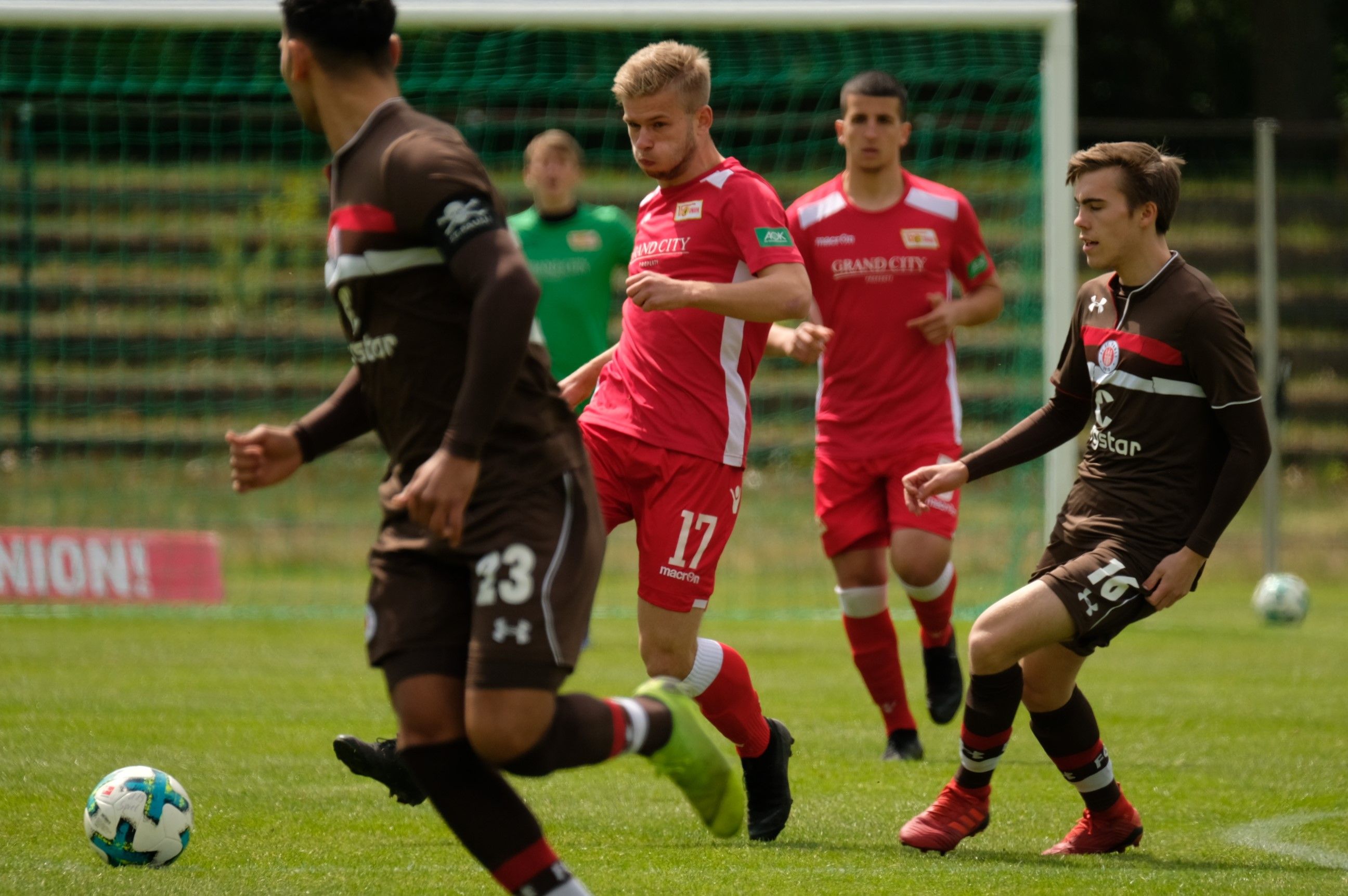 Spielszene eines Fußballspiels mit mehreren Spielern in roten und braunen Trikots auf einem grünen Rasenplatz.