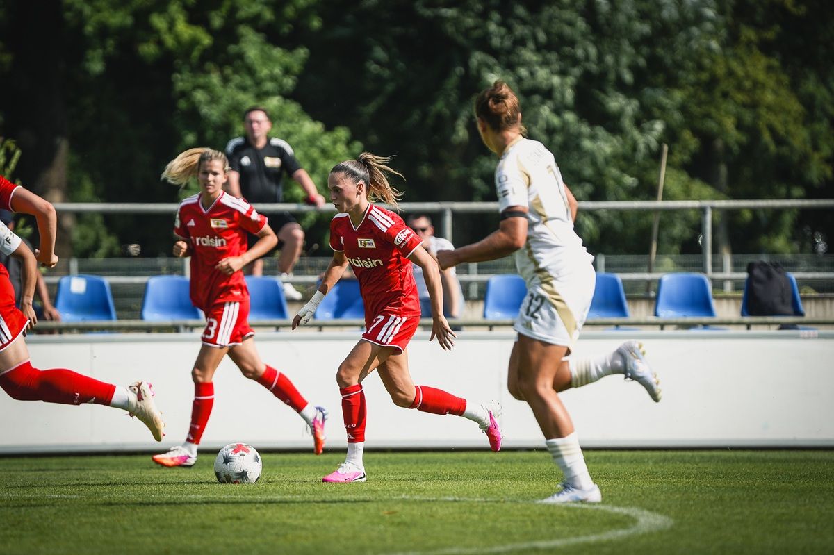 Spielerinnen in rot und weiß auf dem Fußballfeld, im Wettkampf, mit Zuschauerreihen im Hintergrund und einem Schiedsrichter.