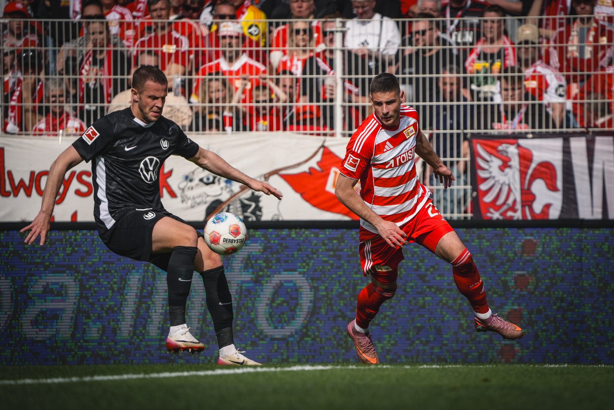 Spielszene aus einem Fußballspiel mit zwei Spielern, einer in schwarzer und einer in roter Uniform, umgeben von jubelndem Publikum.