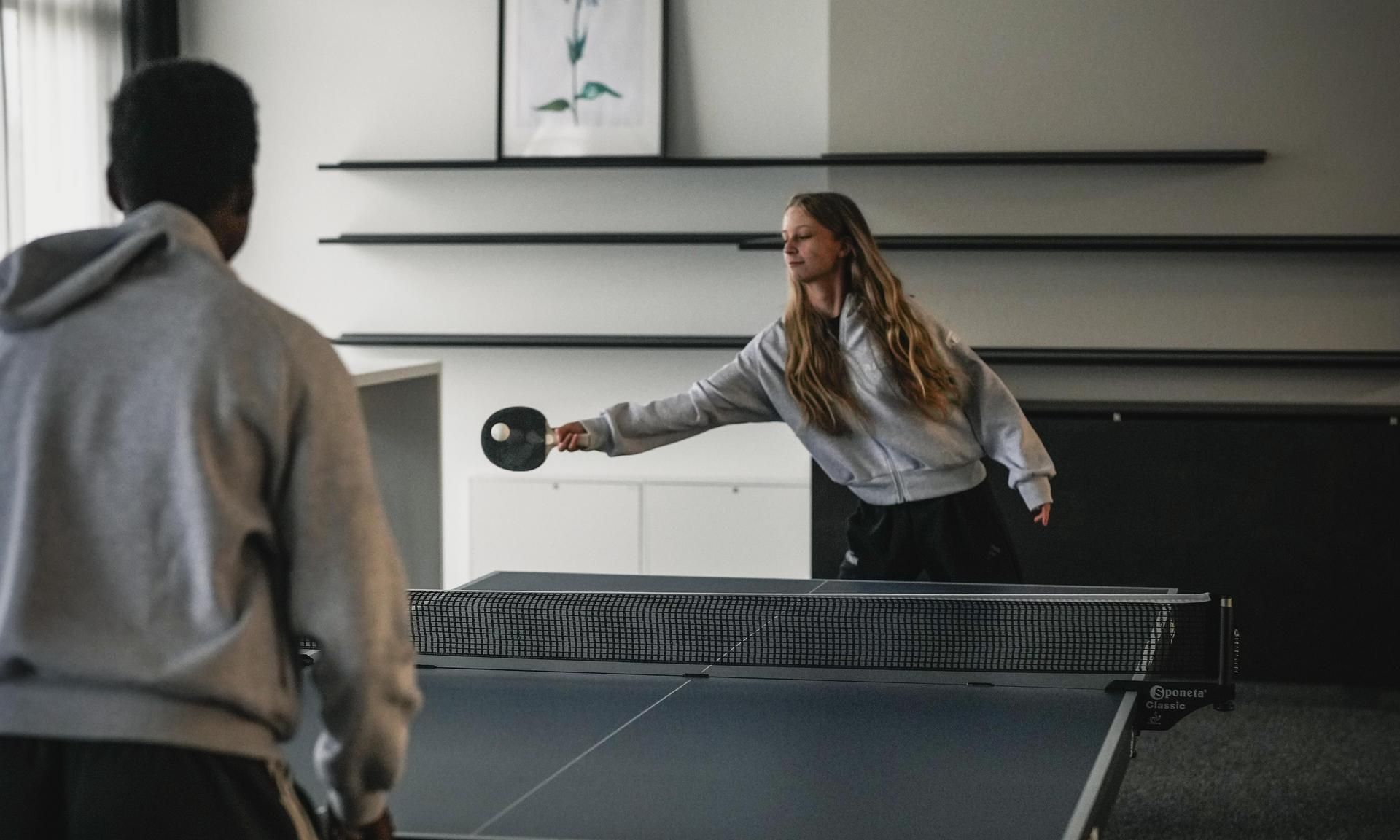 Ein Mädchen mit langen Haaren spielt Tischtennis gegen einen Jungen in einem sportlichen Raum.