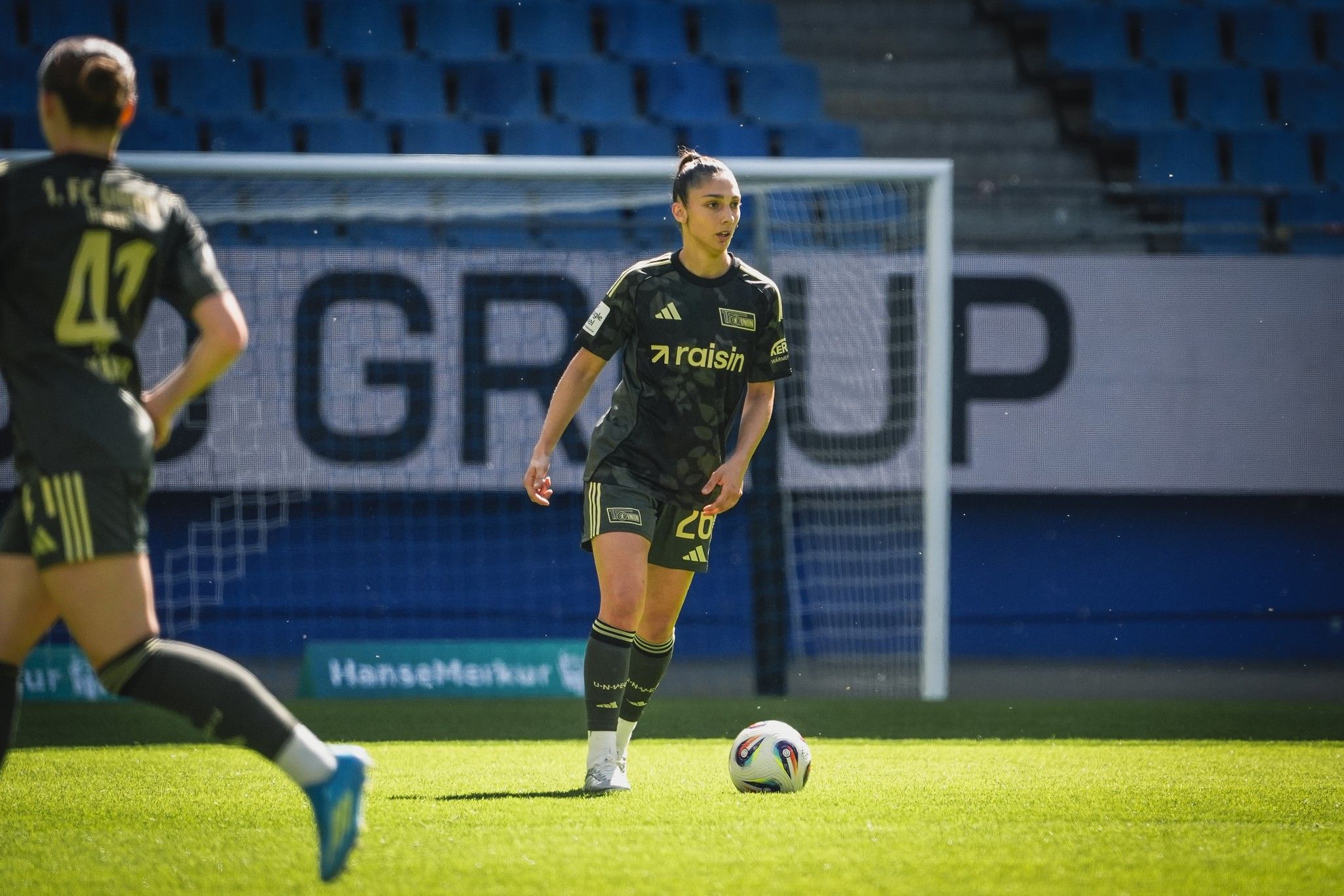 Eine Fußballspielerin mit der Nummer 26 steht bereit, den Ball auf einem grünen Spielfeld zu spielen. Im Hintergrund ist das Tor sichtbar.