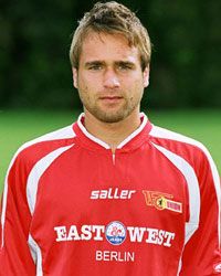 Ein junger Mann in einem roten Fußballtrikot mit dem Logo eines Vereins und der Aufschrift "EAST WEST BERLIN".