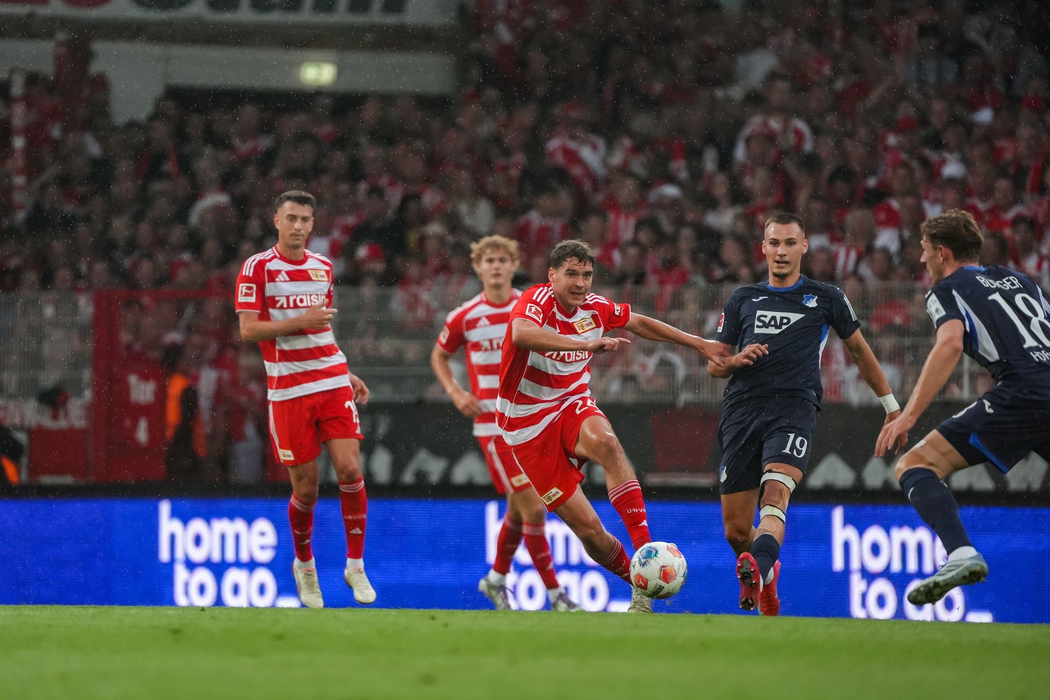 Ein Fußballspieler im roten Trikot passt den Ball, während andere Spieler in blauen Trikots heranlaufen; Publikum im Hintergrund.
