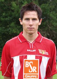 Ein Mann in einem roten Fußballtrikot steht seriös vor einer grünen Kulisse.