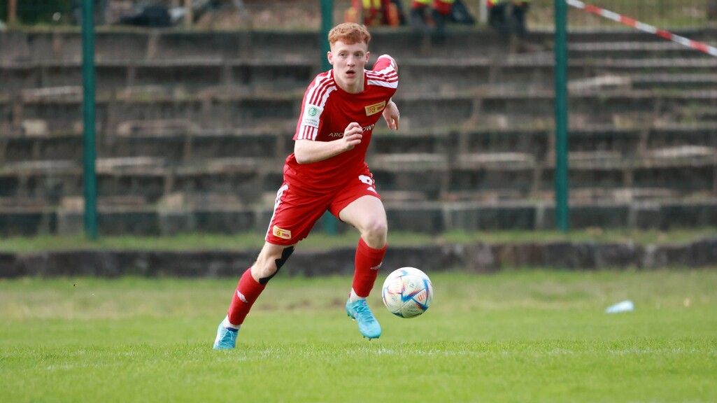 Ein junger Fußballspieler in roter Ausrüstung sprintet über ein Grasfeld, während er den Ball verfolgt.