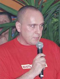 Ein Mann mit Glatze hält ein Mikrofon und spricht, während er in einem Raum mit Pflanzen steht.