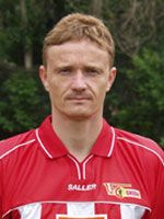 Ein Mann mit kurzer, heller Frisur trägt ein rotes Fußballtrikot und blickt direkt in die Kamera. Hintergrund mit Bäumen.