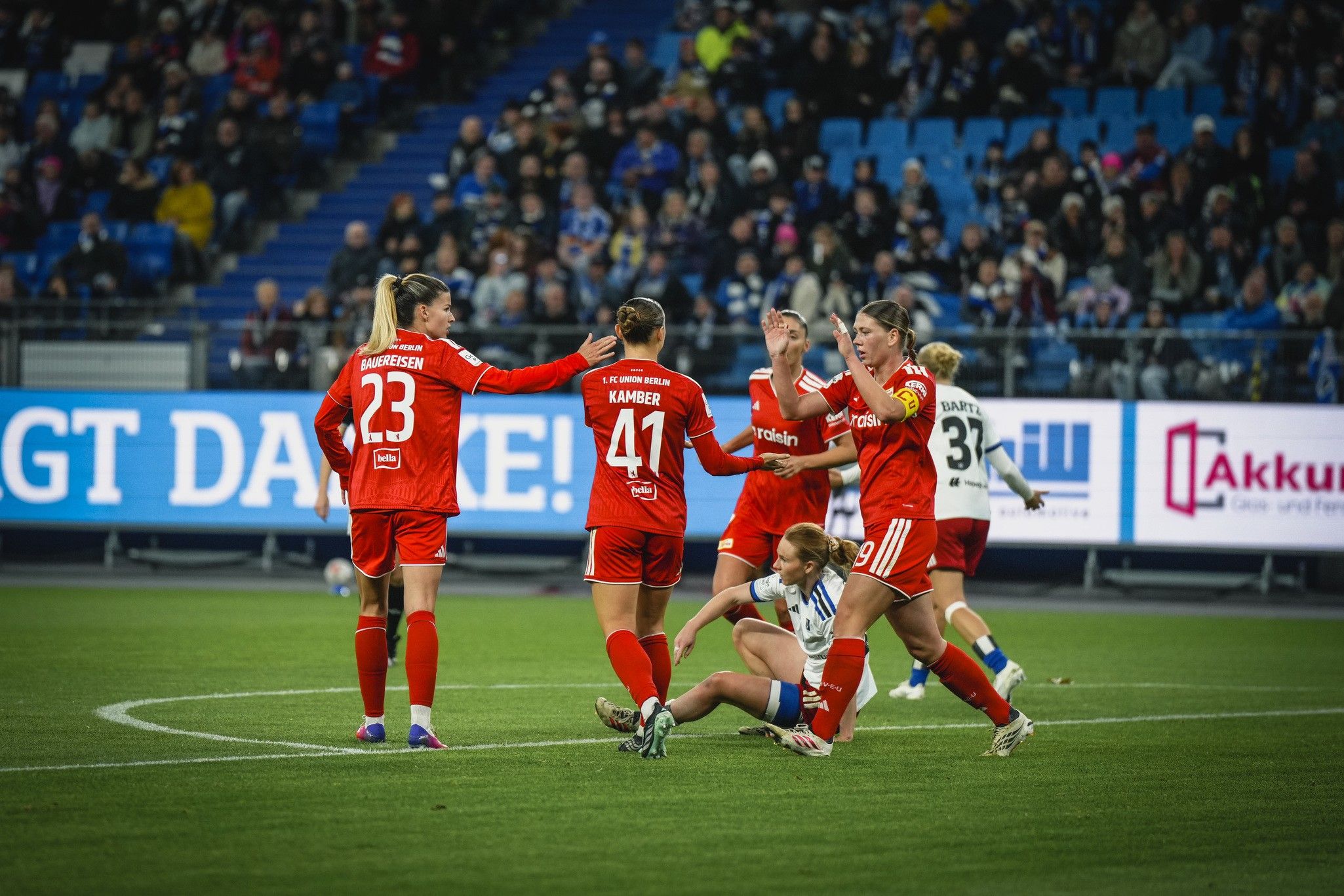 Teammitglieder feiern einen erfolgreichen Spielzug auf dem Fußballfeld, während eine Spielerin am Boden liegt.