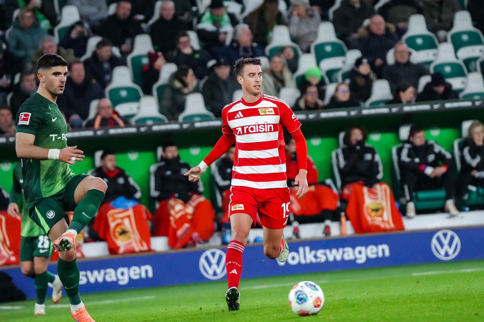 Ein Fußballspieler in rotem Trikot dribbelt den Ball, während ein Gegner in grünem Trikot bereit ist, zu reagieren. Fans im Hintergrund.