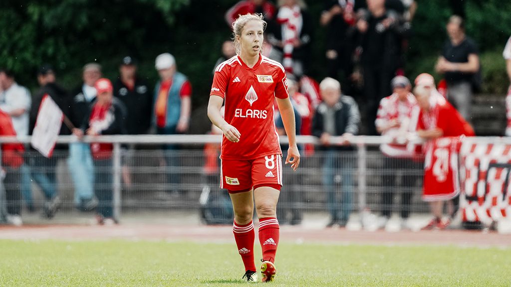A female soccer player in a red jersey with the inscription "ELARIS" runs on the field, with fans in the background.