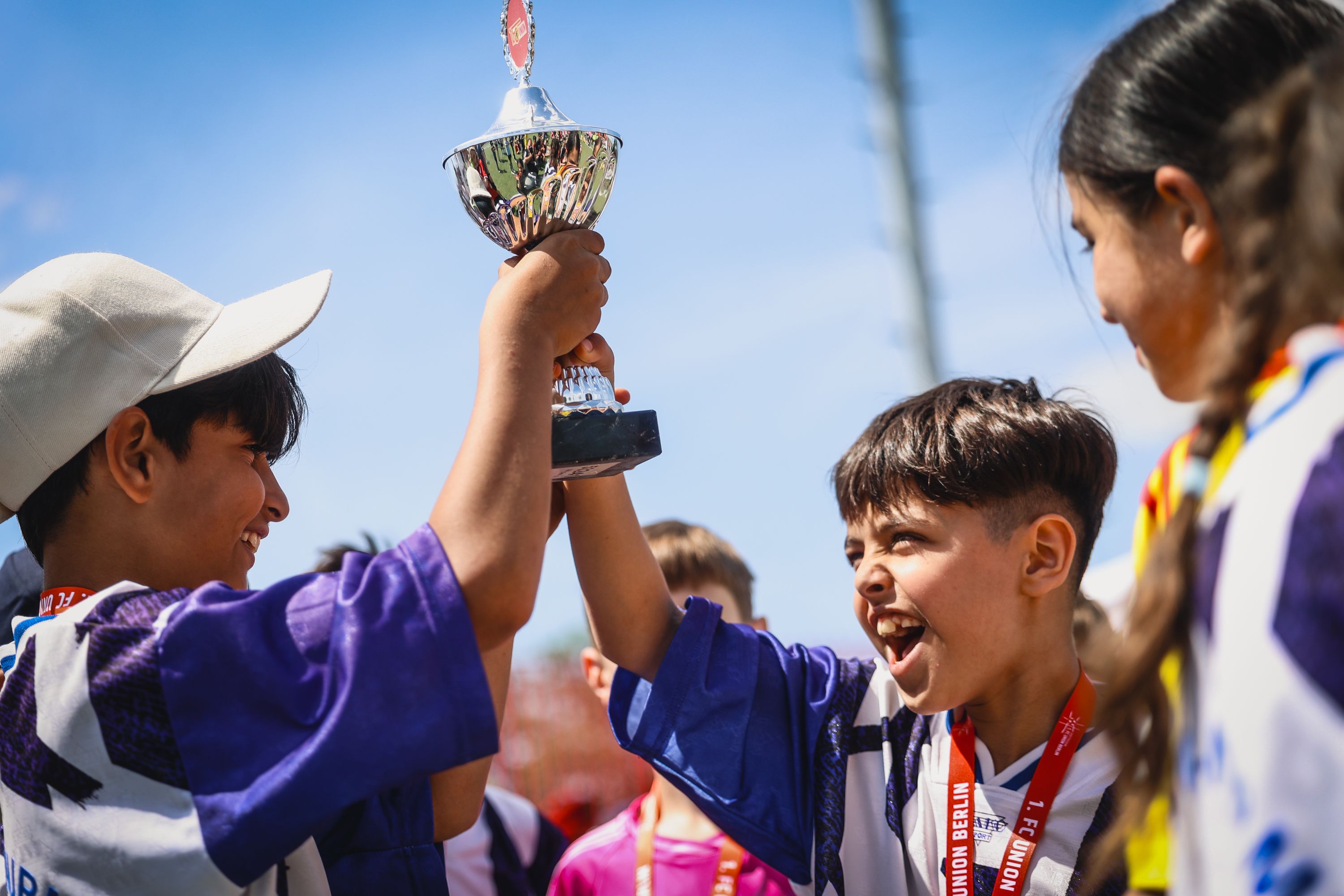 Zwei Kinder jubeln und halten gemeinsam einen Pokal hoch, umgeben von anderen fröhlichen Kindern.