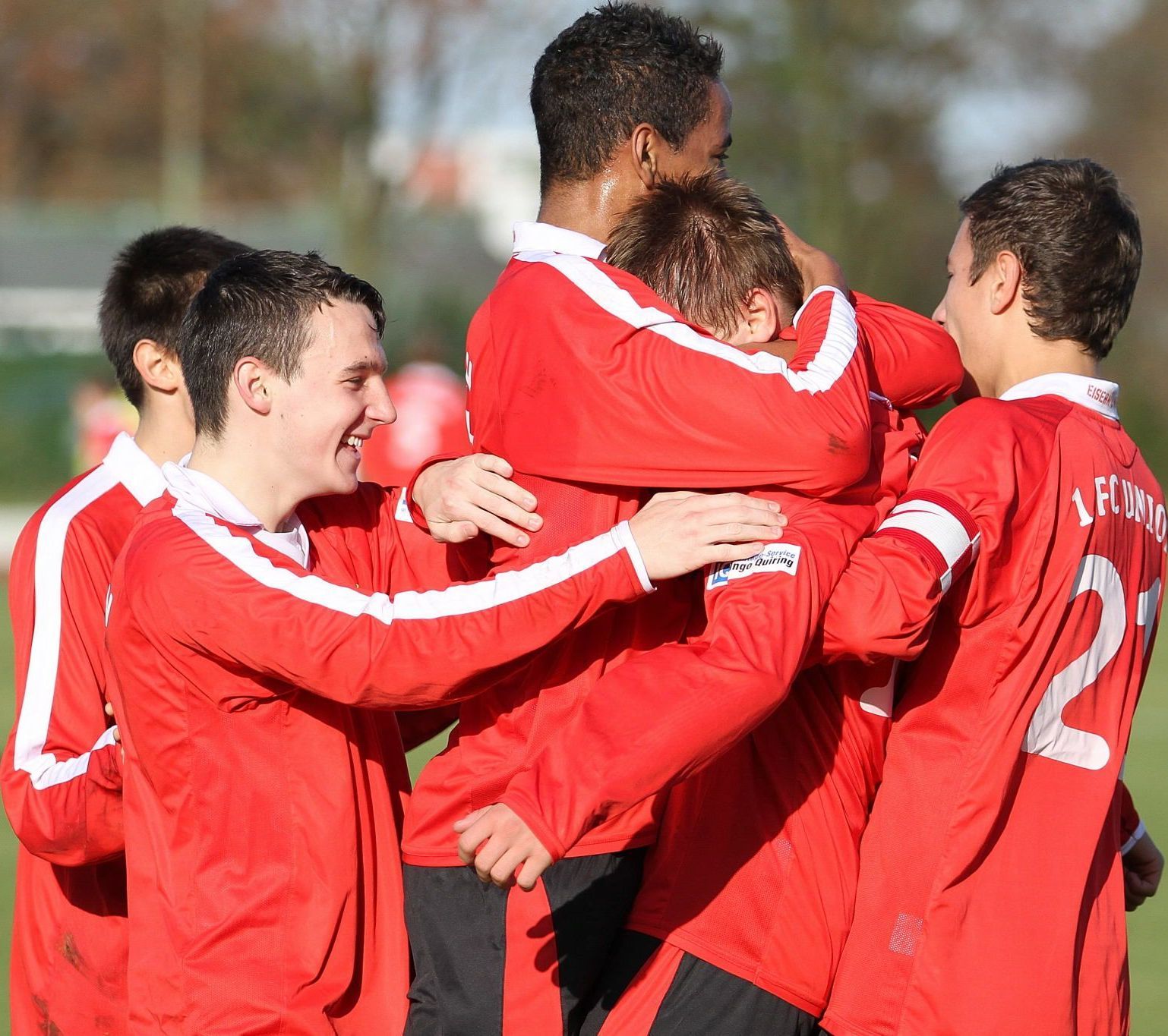 Sechs junge Fußballspieler in roten Trikots umarmen sich nach einem Tor. Freude und Teamgeist sind deutlich zu sehen.