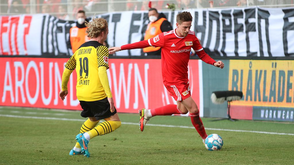 A player in red club gear dribbles the ball while an opposing player in yellow clothes follows him.