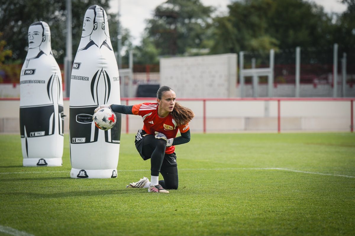 Eine Fußballtorhüterin kniet auf dem Rasen und hält einen Ball in der Hand, während Trainingshindernisse im Hintergrund stehen.