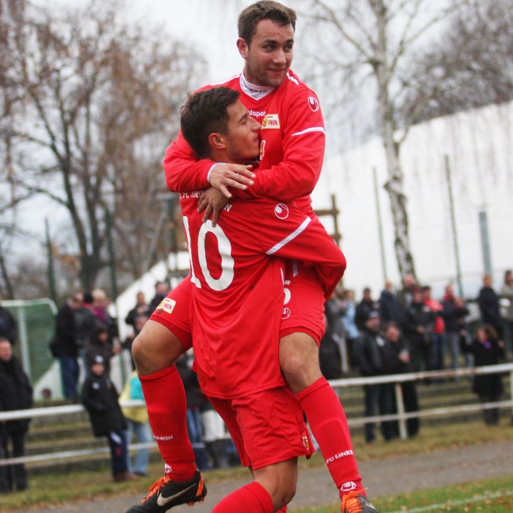 Zwei Fußballspieler in roten Trikots feiern zusammen auf dem Platz, einer hebt den anderen auf den Schultern.