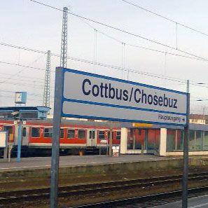 Ein Bahnhofsschild mit dem Schriftzug "Cottbus/Chosebuz" im Vordergrund, im Hintergrund eine Lokomotive und Bahnhofseinrichtungen.