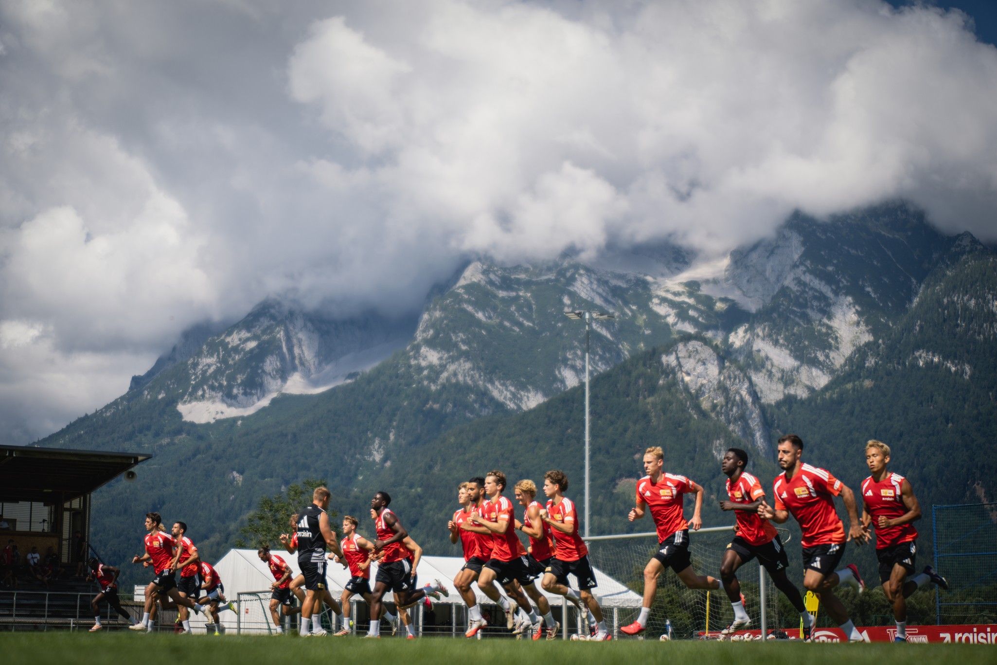 Eine Gruppe von Athleten in roten Trikots joggt auf einem Sportfeld vor einer beeindruckenden Bergkulisse unter einem bewölkten Himmel.