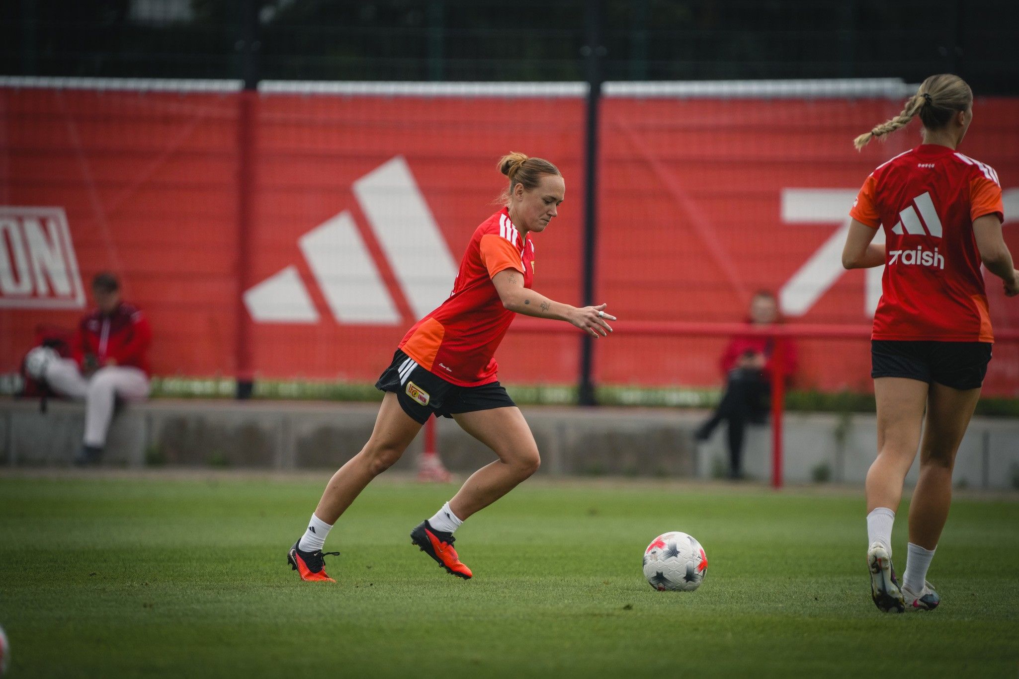 Eine Fußballspielerin in orangefarbener Kleidung trainiert auf einem grünen Platz mit einem Ball. Im Hintergrund sind rote Netzplanen zu sehen.