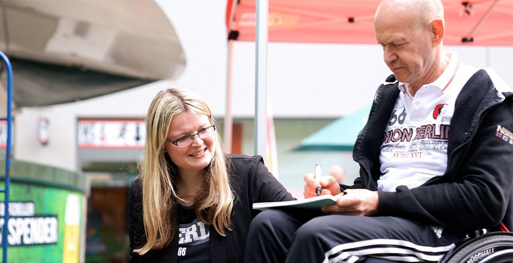 Eine Frau sitzt neben einem mann in einem Rollstuhl, während dieser in ein Notizbuch schreibt. Beide lächeln.