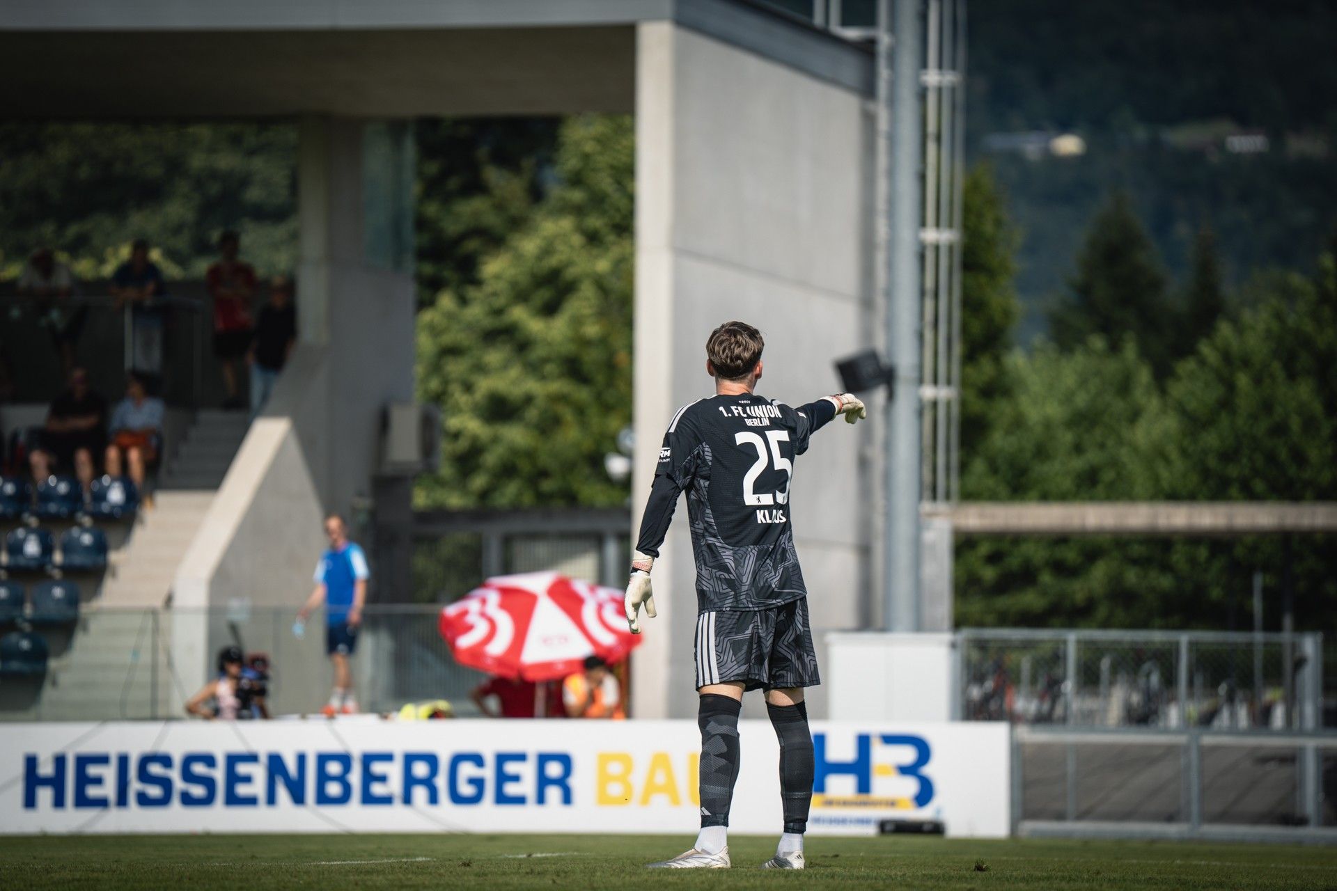 Ein Torwart in schwarzer Ausrüstung zeigt auf das Spielfeld, während er vor einer Tribüne mit Zuschauern steht.