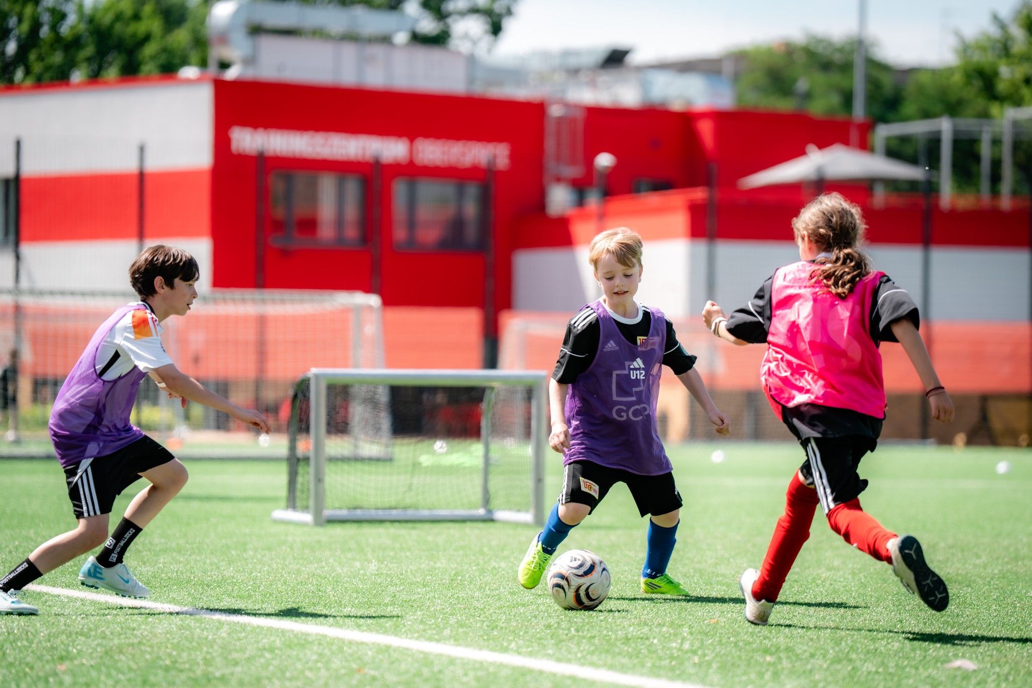 Drei Kinder spielen Fußball auf einem grünen Platz, während ein Tor im Hintergrund zu sehen ist.