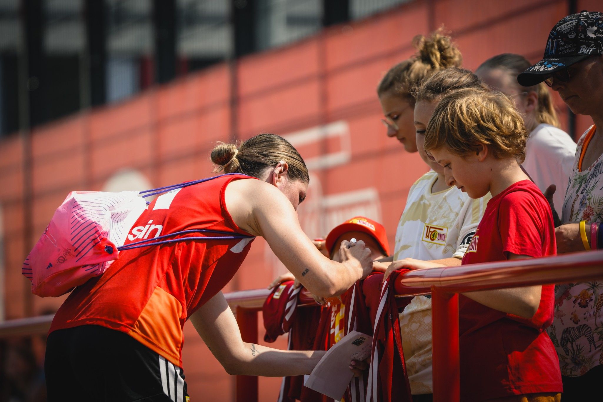 Eine Sportlerin in rot signiert Autogramme für Fans, darunter ein Junge mit rotem Hut, im Hintergrund weitere Zuschauer.