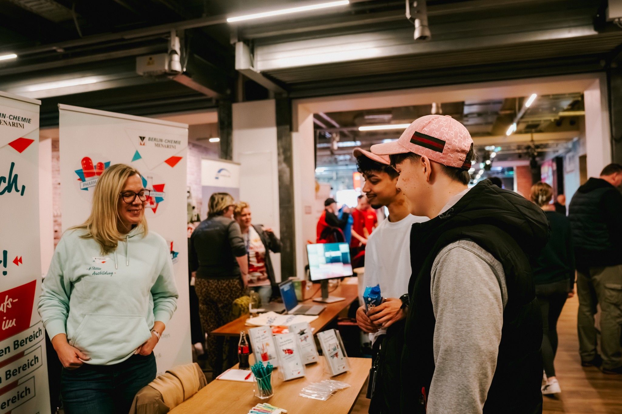 Zwei junge Männer unterhalten sich mit einer Frau an einem Messestand, umgeben von Informationsmaterial und Besuchern.