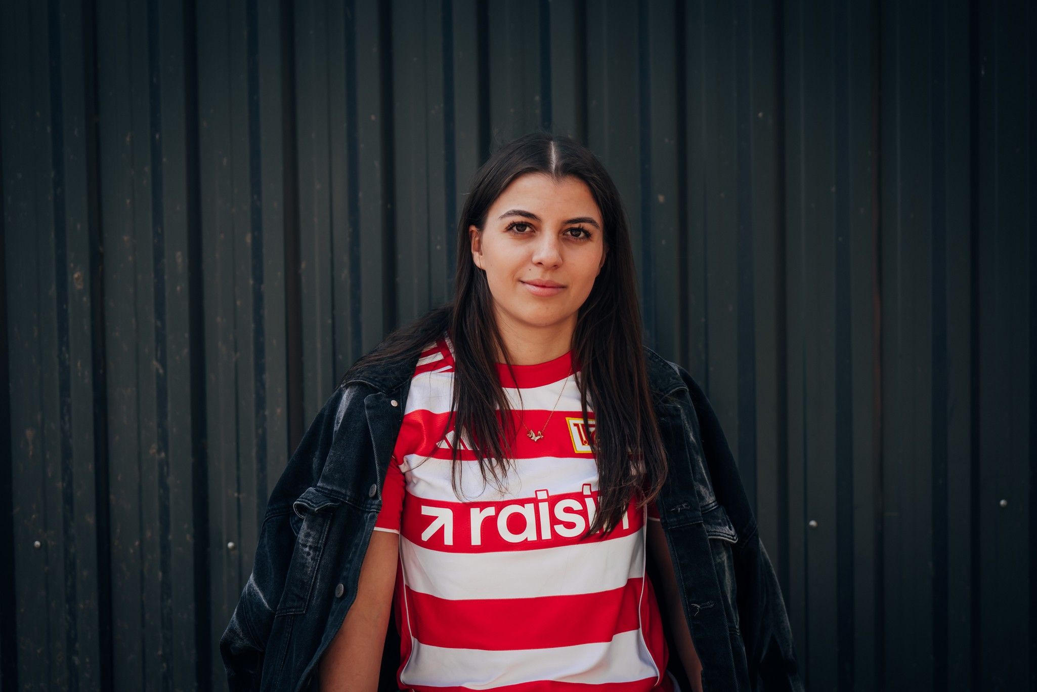 Eine junge Frau mit langen, dunklen Haaren trägt ein rot-weiß gestreiftes Trikot und eine schwarze Jacke vor einem grafischen Hintergrund.