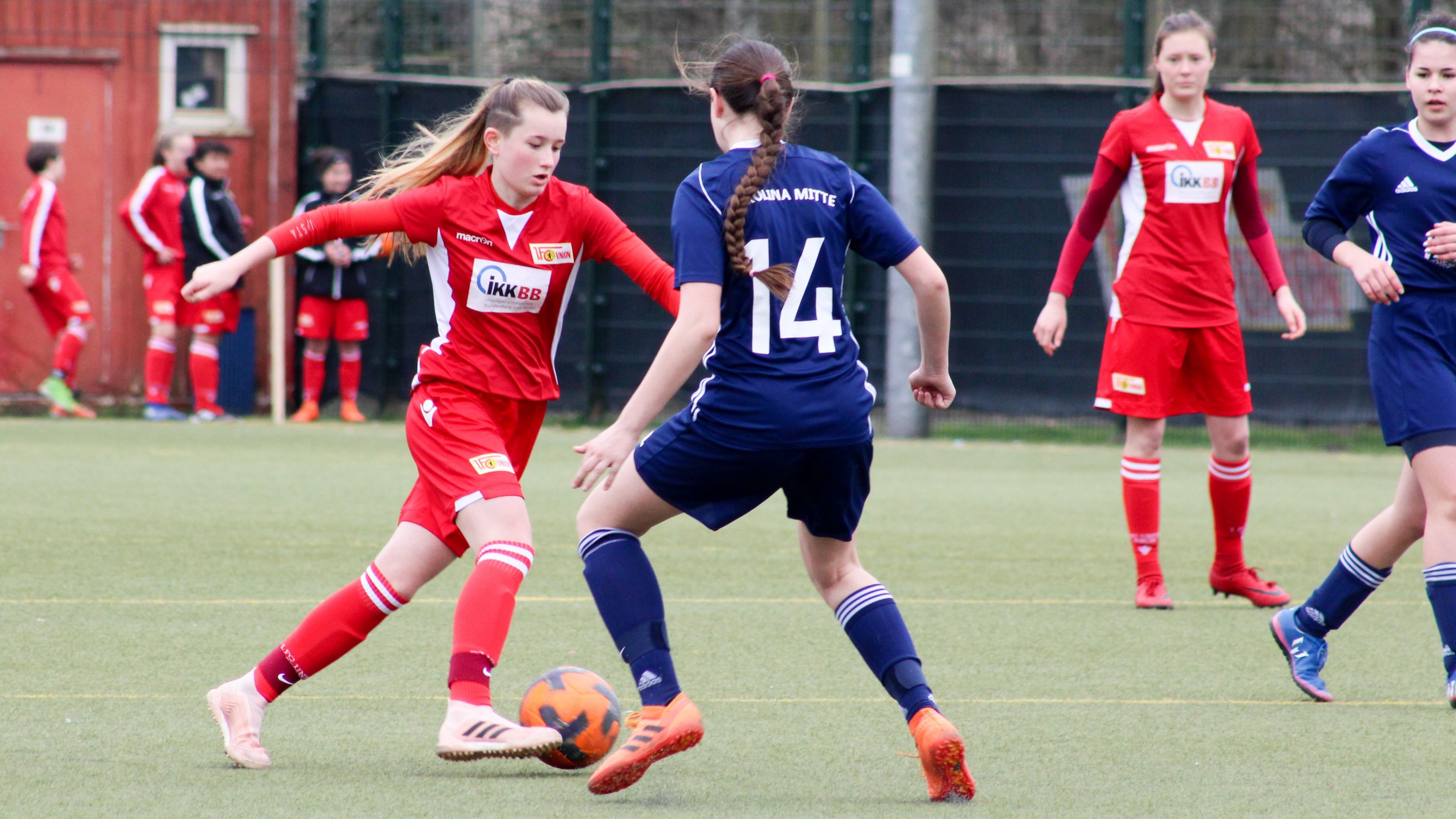 Zwei Fußballspielerinnen im Wettkampf um den Ball auf einem Kunstrasenplatz. Eine Spielerin trägt ein rotes Trikot, die andere ein blaues.