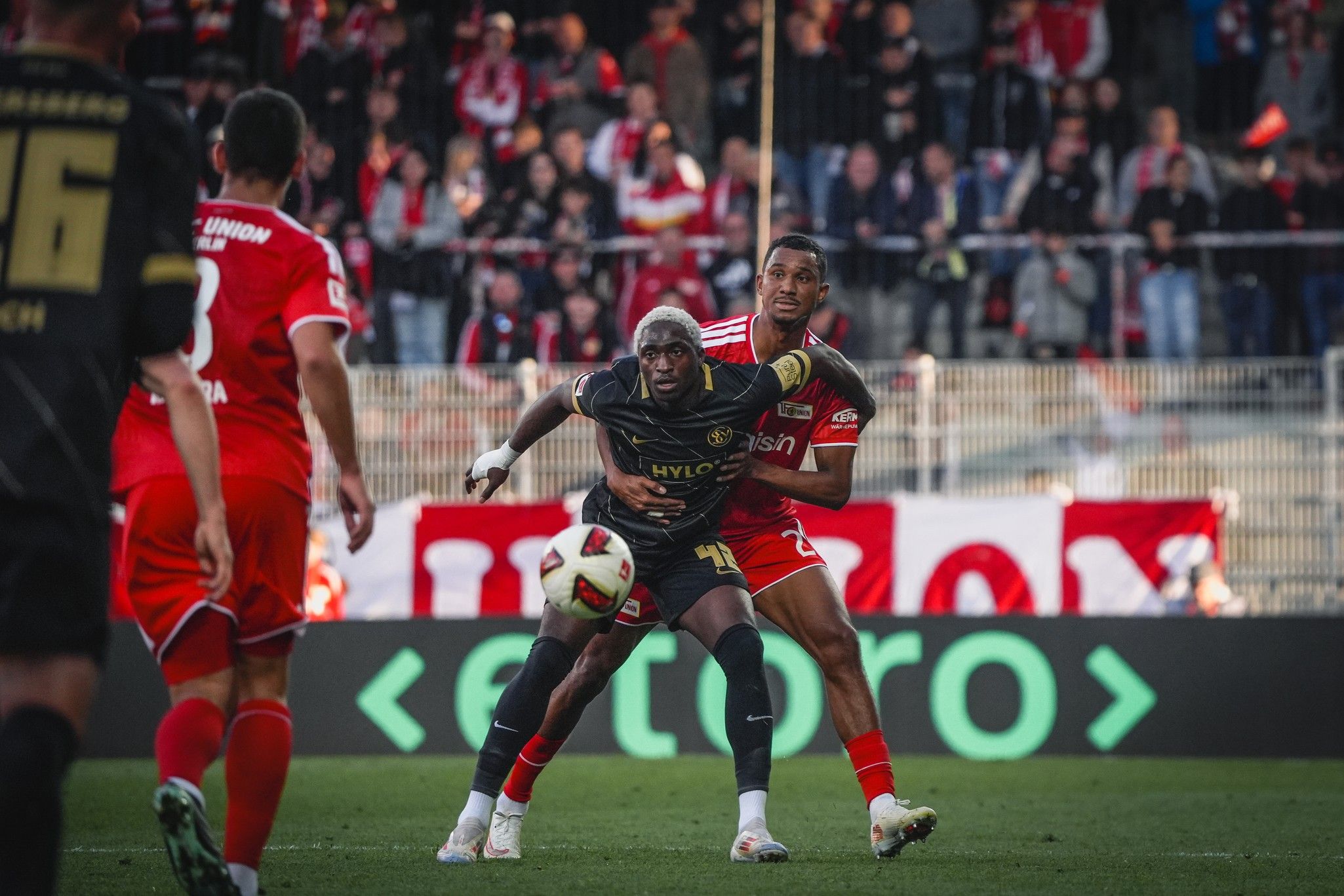 Zwei Fußballspieler kämpfen um den Ball, während Zuschauer im Hintergrund jubeln. Stadionatmosphäre während eines Spiels.