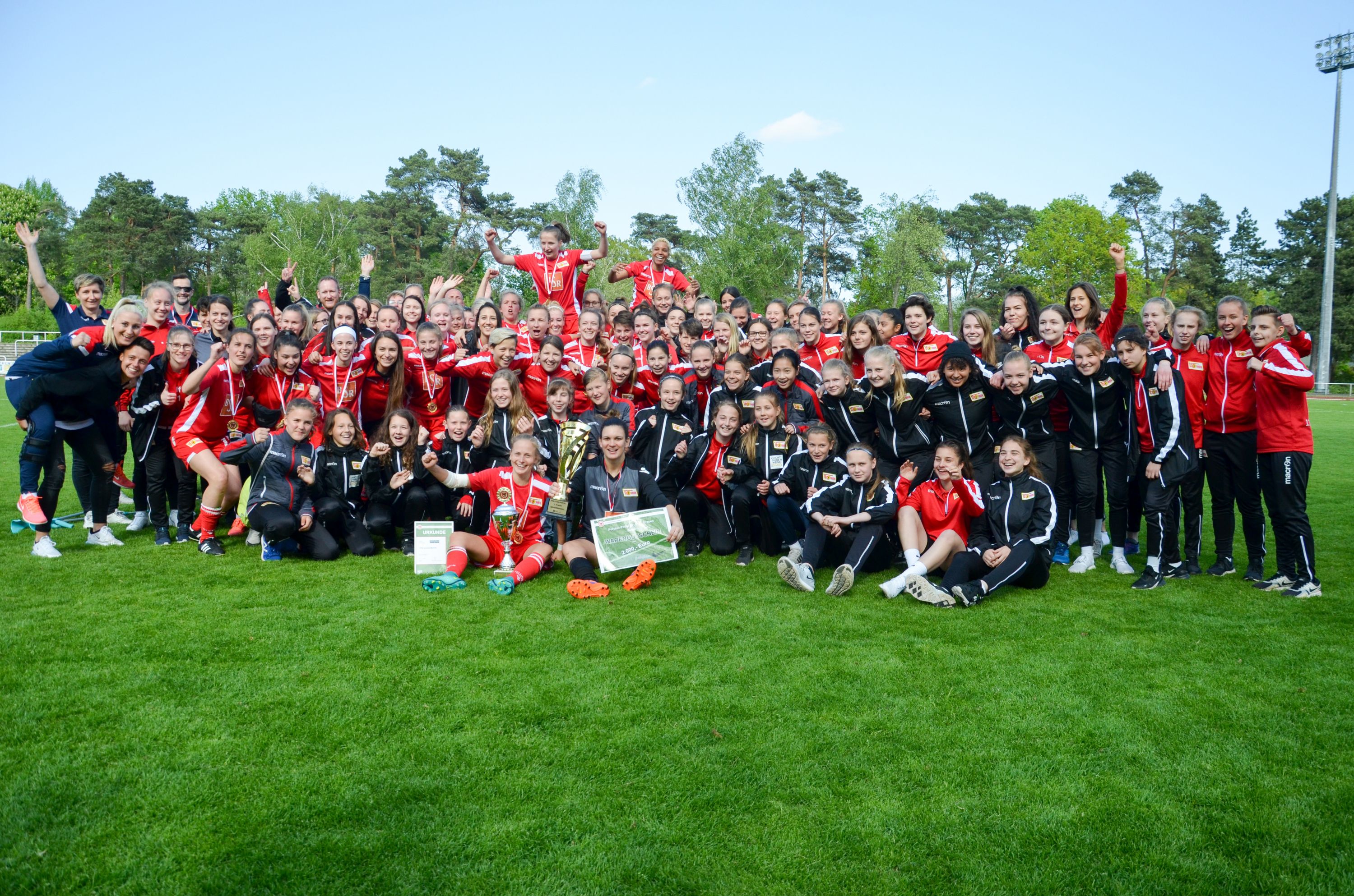 Gruppenshot von vielen Personen in Sportkleidung, die jubelnd auf einem Fußballfeld stehen, mit Pokal in der Mitte.