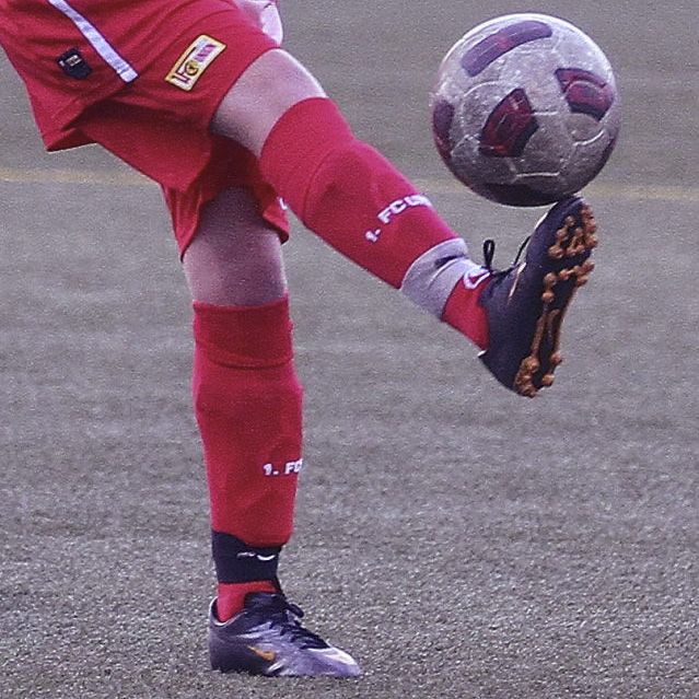 Ein Fußballspieler in roter Ausrüstung dribbelt einen Fußball auf einem Sportplatz.