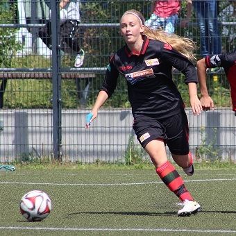 Eine Fußballspielerin in schwarzem Trikot sprintet mit dem Ball auf einem Rasenplatz. Zuschauer sind im Hintergrund sichtbar.
