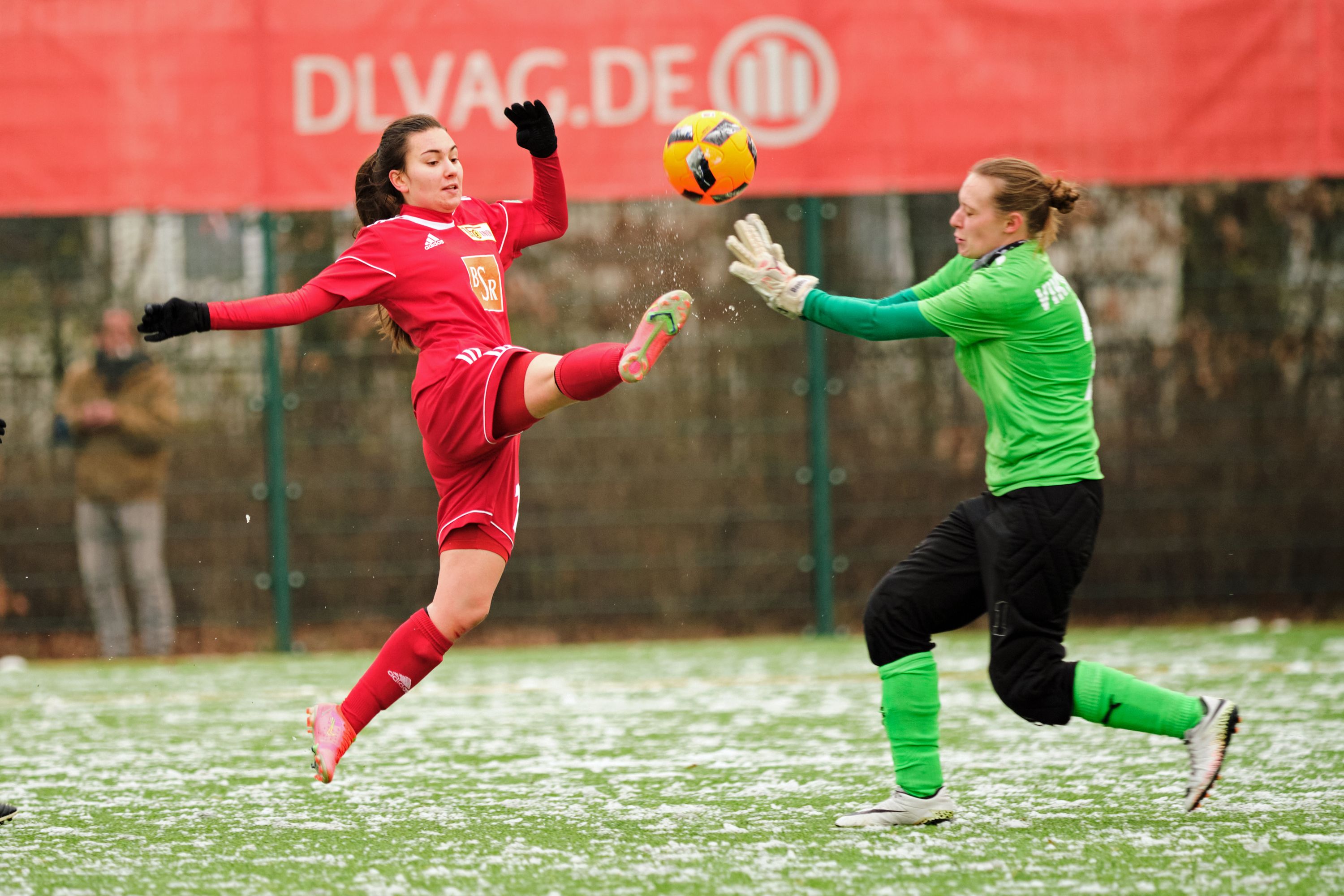 Spielszene im Fußball: Eine Spielerin in rotem Trikot hebt das Bein, während die Torhüterin in grün zum Ball greift.