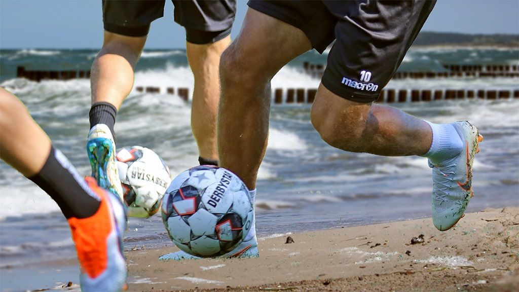 Two players are dribbling a soccer ball on the beach, with waves in the background and a ball on the sand.