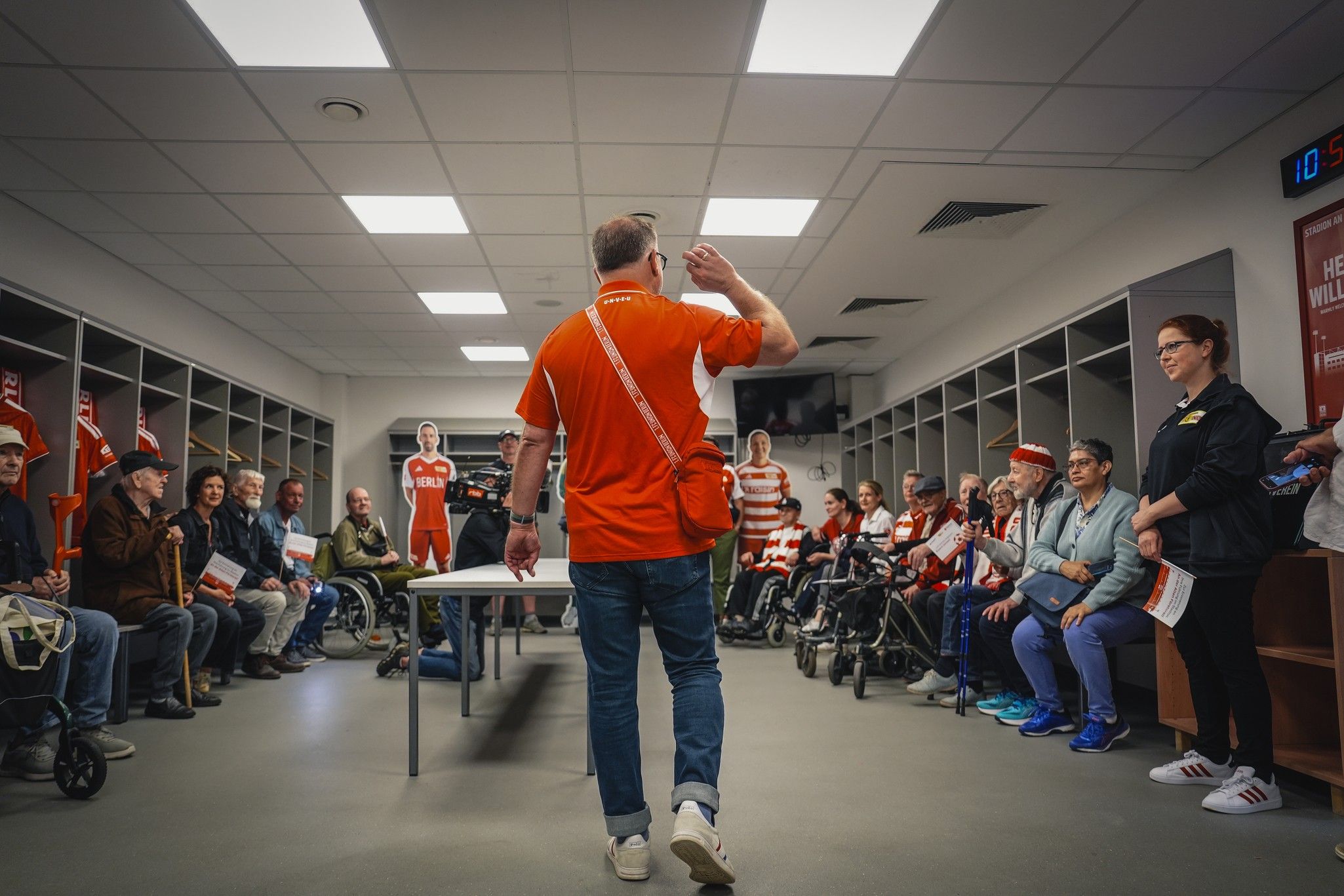 Ein Mann in rotem Shirt steht vor einer Gruppe von Menschen in einem Raum mit Tischen und Stühlen, während sie aufmerksam zuhören.