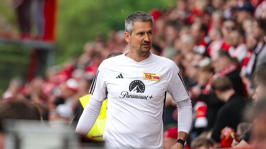 Ein Trainer in einem weißen Vereins-T-Shirt steht auf dem Spielfeld, umgeben von jubelnden Fans im Hintergrund.